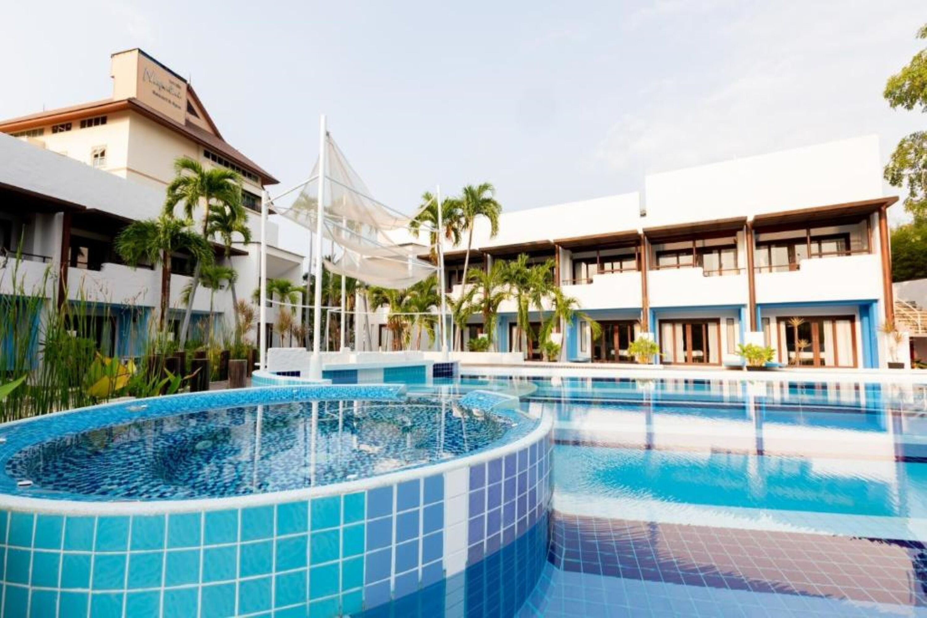 A hotel with a large swimming pool, palm trees, and a modern building with multiple balconies.