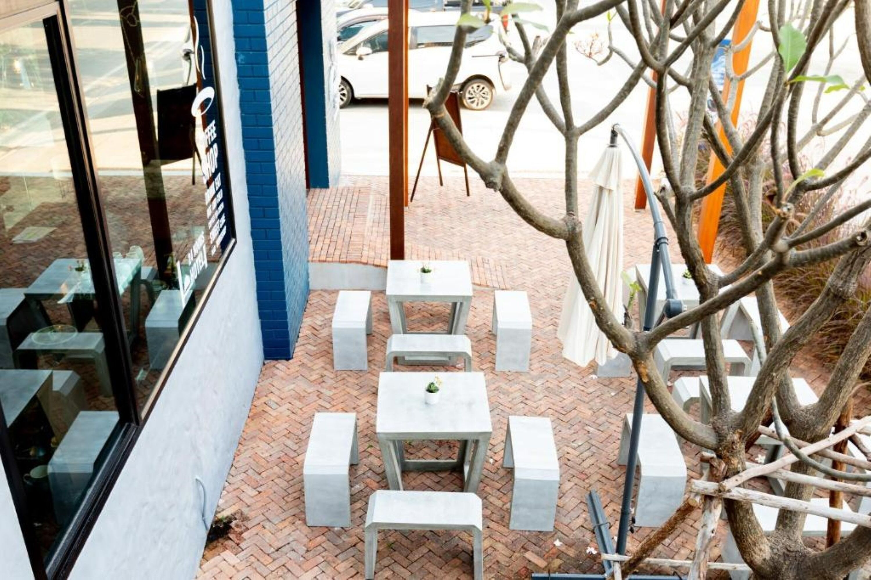 A patio area with white tables and benches, brick flooring, and a view of the street.