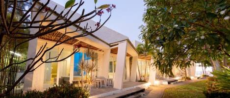 A modern house with a covered patio, a well-maintained garden, and a clear view of the interior through large windows.