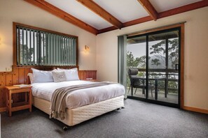 A bedroom with a large bed, wooden headboard, a nightstand, a wall-mounted light, and a sliding glass door leading to an outdoor area with a chair and table. - Freycinet Cottage 2 - Freycinet Holiday Houses (Coles Bay)