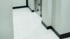 Un corridoio con pavimento piastrellato di bianco, una porta di vetro per l'ascensore e un allarme di emergenza fissato al muro.