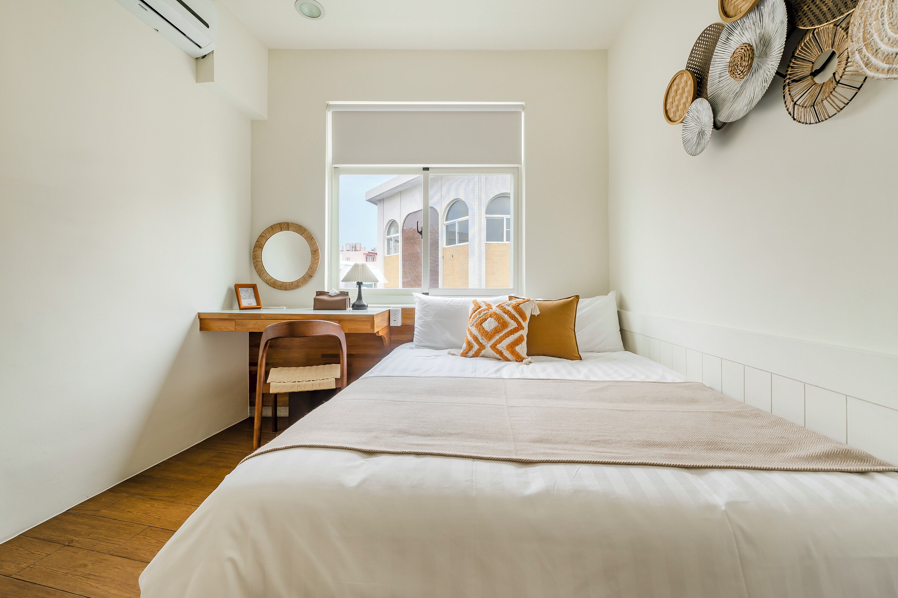 A bedroom with a bed, a desk, a chair, a window, and a view of buildings outside.