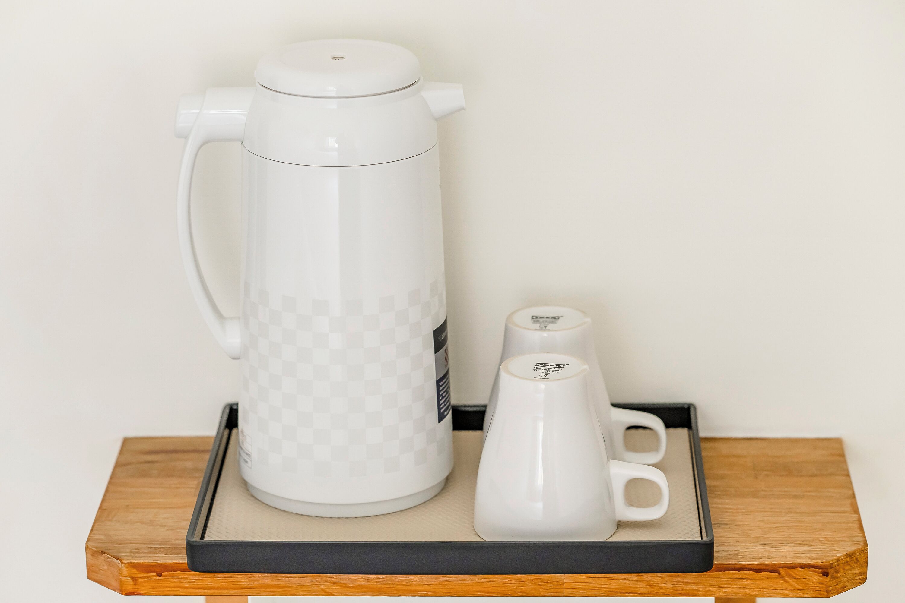 Een witte koffiezetapparaat met een geruit patroon, een witte mok en een dienblad op een houten tafel tegen een effen muur.
