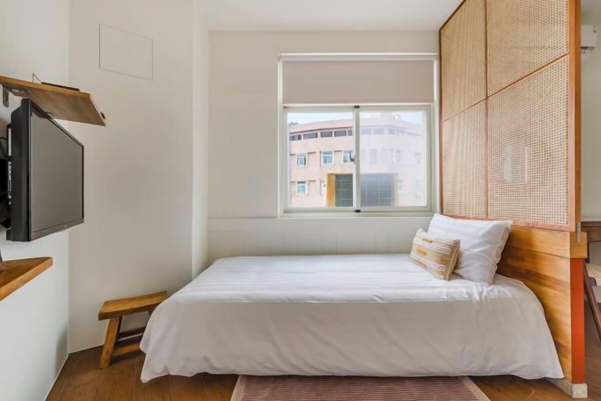 A single bed with a wooden headboard, a television mounted on the wall, and a window with a view of buildings.