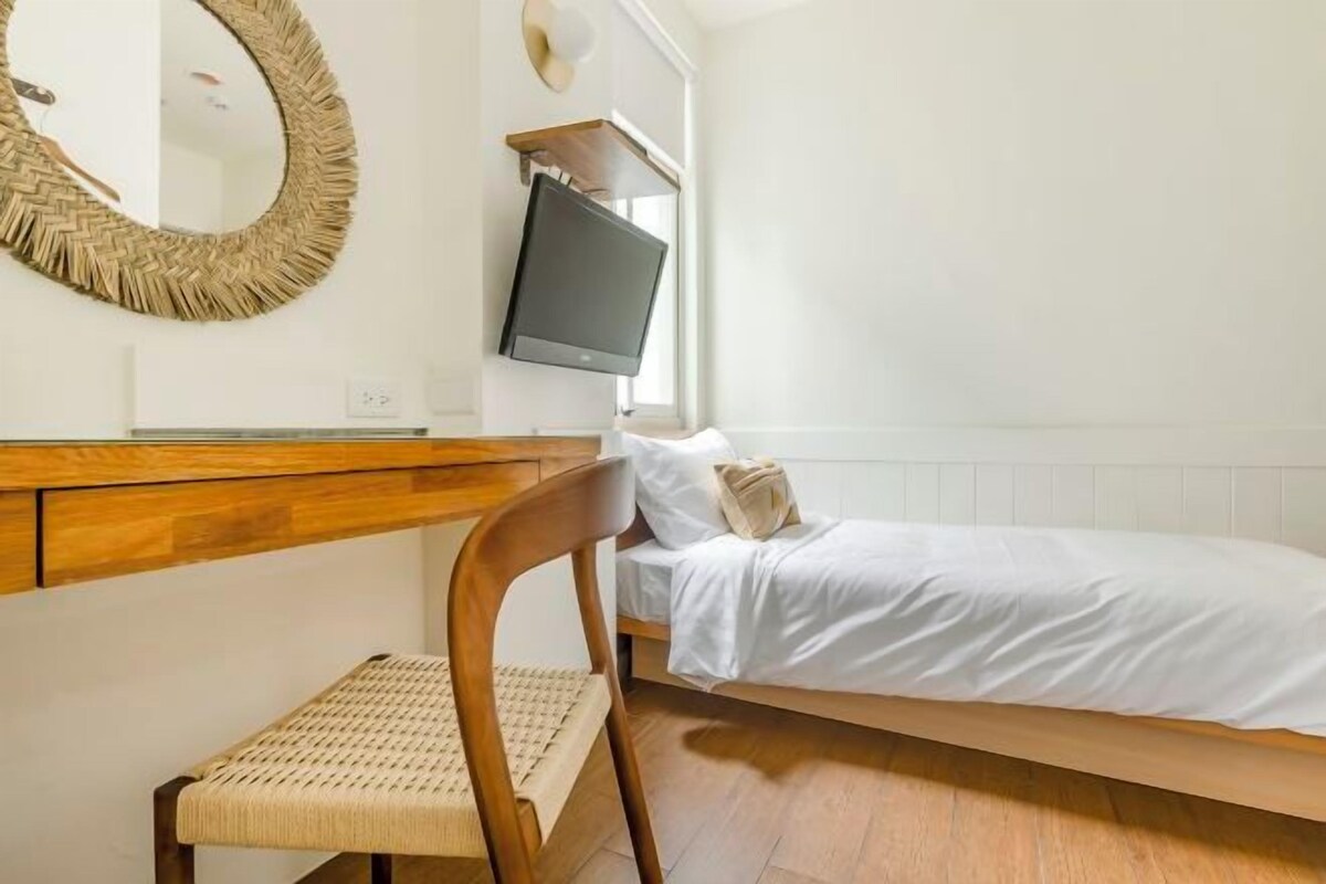 A bedroom with a wooden headboard, a woven chair, a bed with white bedding, a round woven mirror, and a flat-screen TV mounted on the wall.