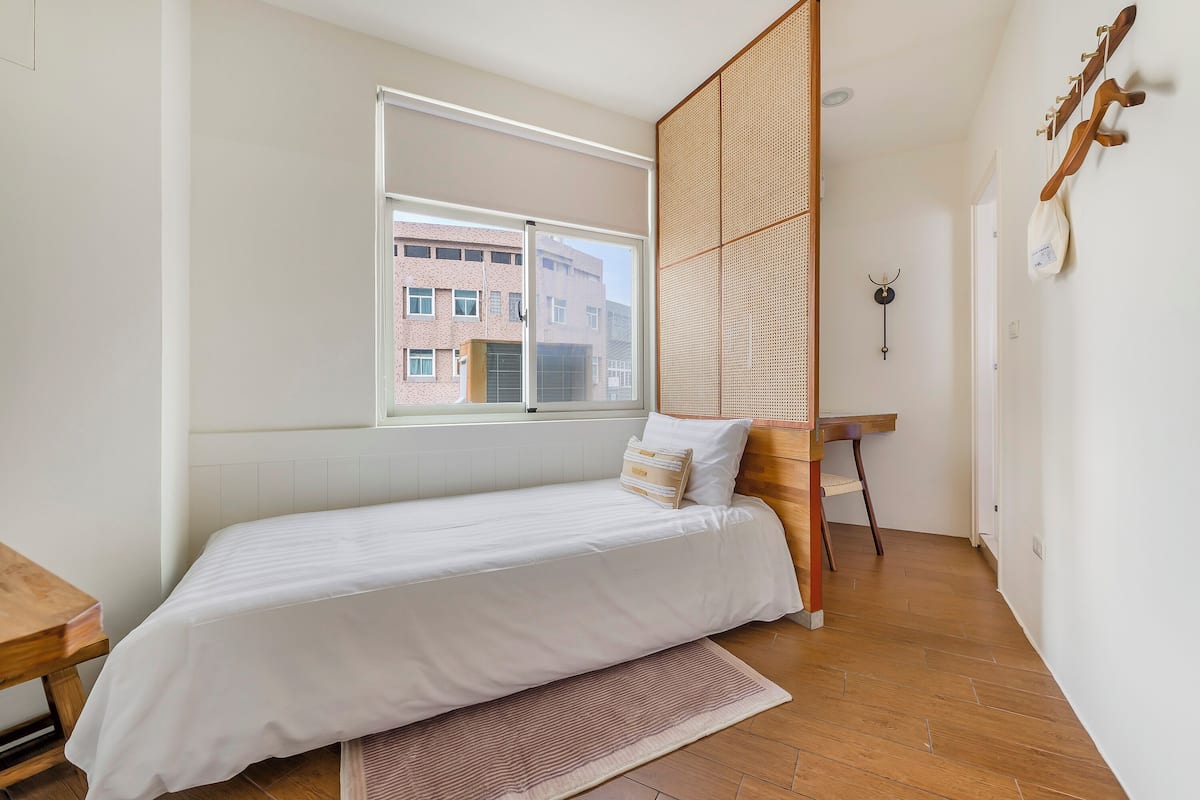 A bedroom with a single bed, a window, a wooden floor, and a view of a building outside.
