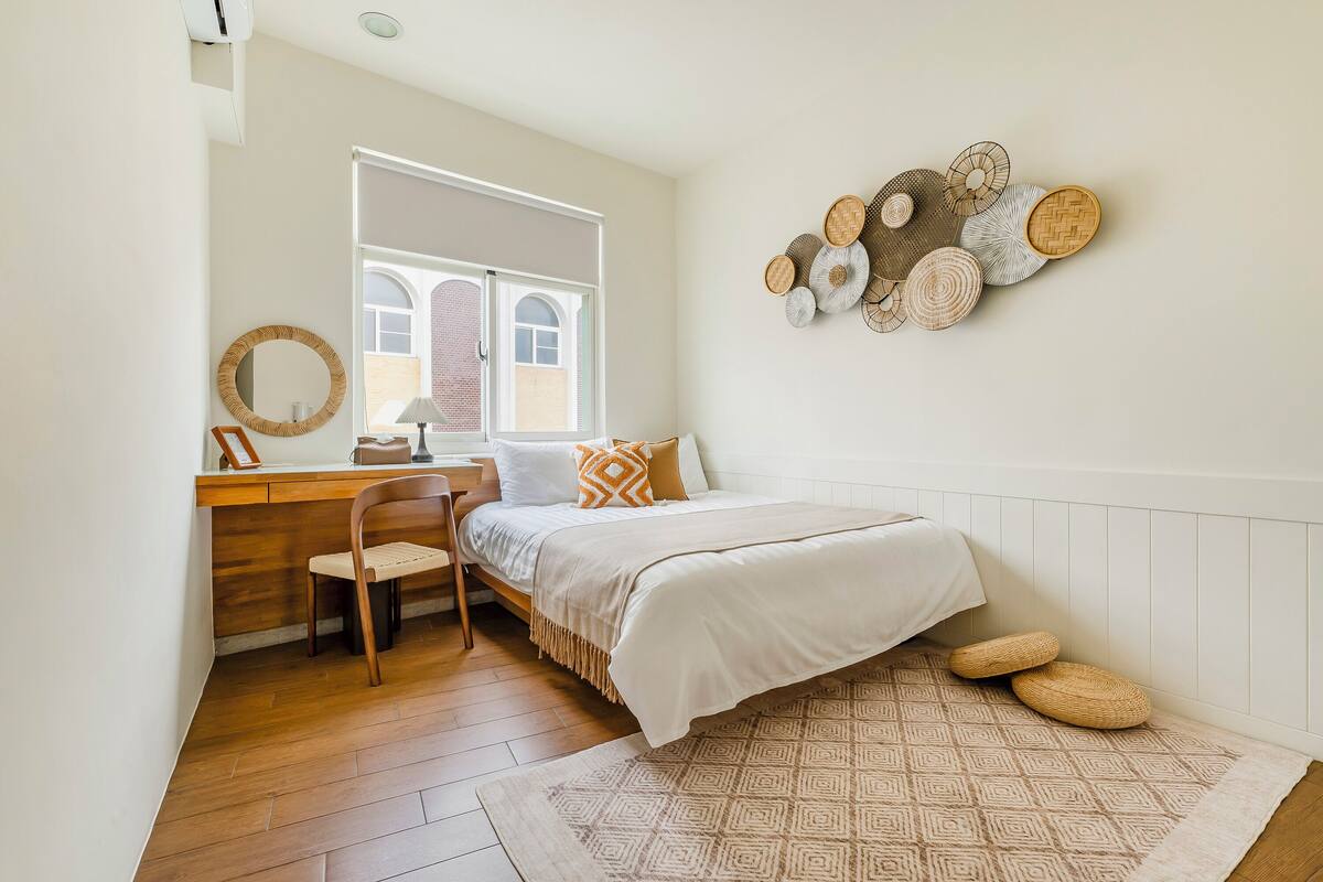 A bedroom with a wooden desk, a chair, a bed with a patterned rug, and a wall with woven wall art.