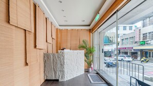 A reception area with a wooden reception desk, a potted plant, and a view of the street outside. - Chin Style Easy Stay (Magong)