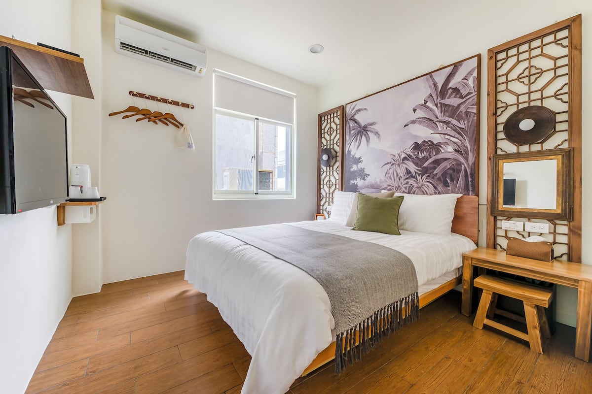 A bedroom with a bed, wooden floor, a window, a wall-mounted air conditioner, a coat rack, and a decorative panel with a palm tree painting.