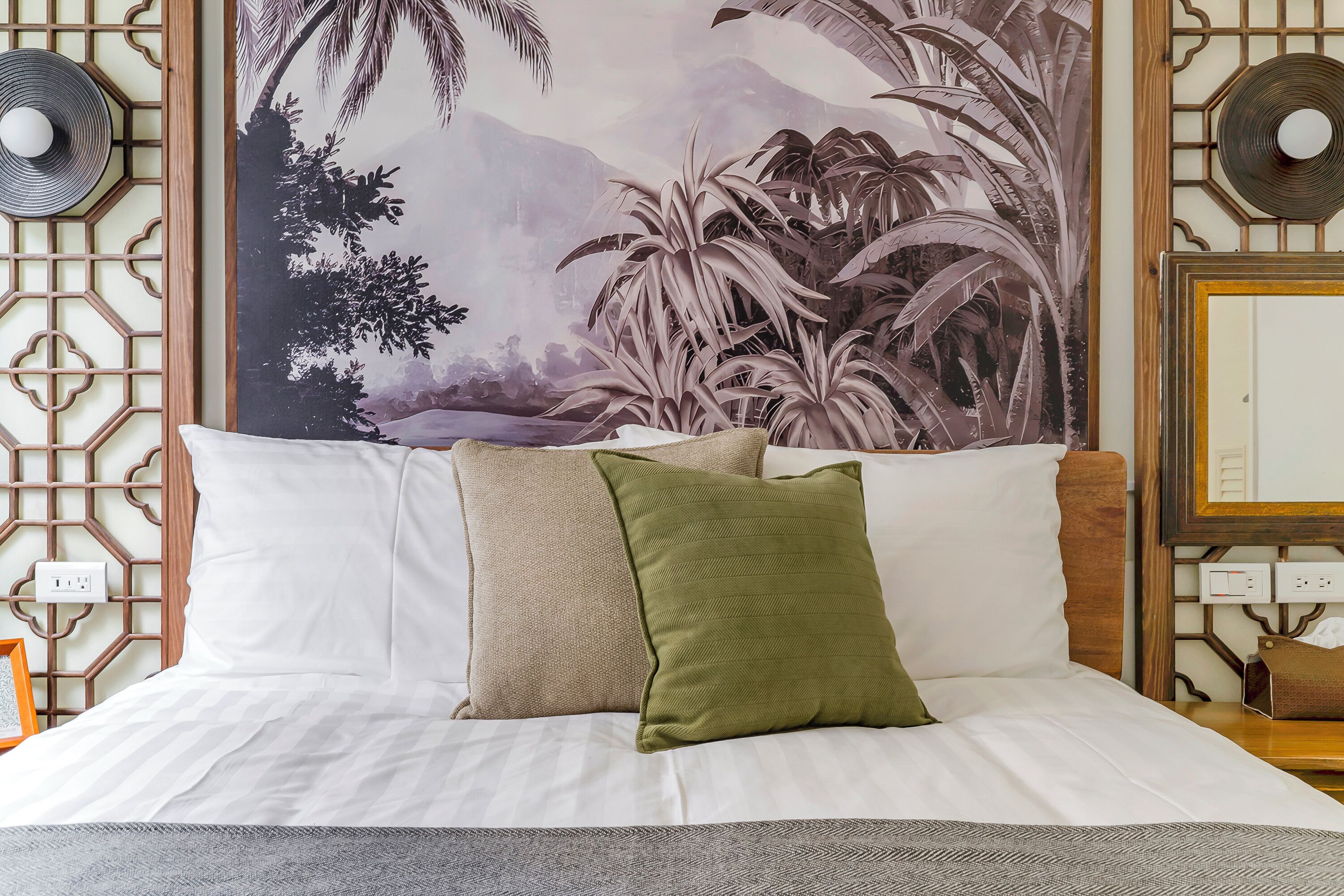 A neatly made bed with white and green pillows, a large tropical mural on the wall, and a traditional wooden partition with a geometric pattern.
