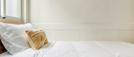 Una cama bien hecha con ropa de cama blanca y una almohada beige. La habitación tiene un cabecero blanco con paneles y una ventana con persianas.