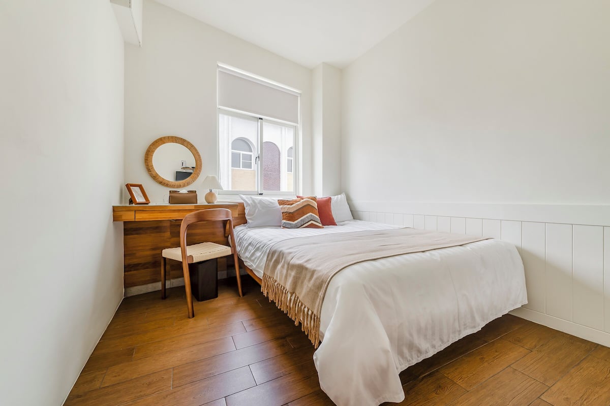 A bedroom with a bed, a wooden desk, a chair, a mirror, and a window.