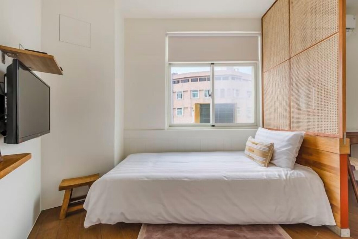 A bedroom with a bed, a window, a television, and a wooden bench.