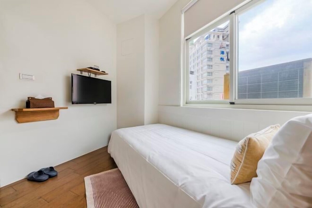 Een slaapkamer met een bed, een aan de muur bevestigde televisie, een raam met uitzicht op gebouwen en een houten vloer.