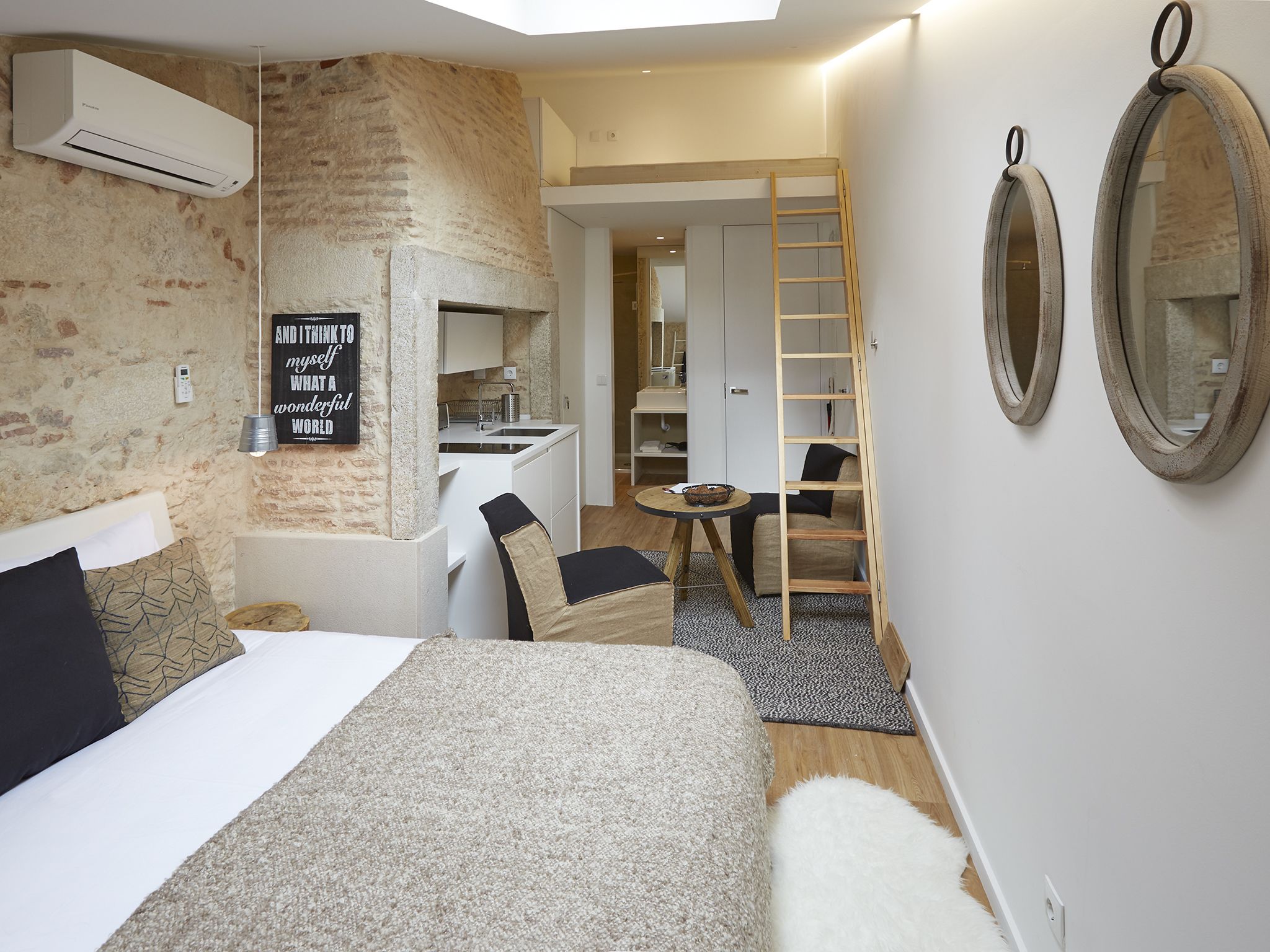 Une chambre d’hôtel compacte comprenant un lit, une petite kitchenette, un coin salon avec une échelle et un miroir au mur.