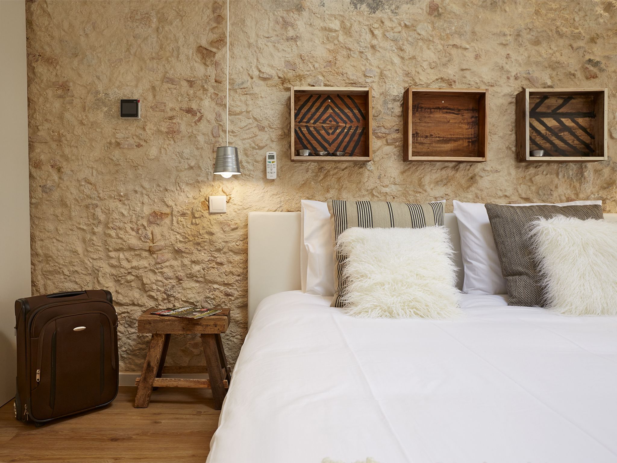 A bedroom with a stone wall, a bed with white bedding, a suitcase, and a nightstand with a lamp and a remote control.