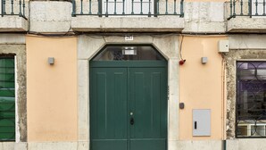 A green double door with a small window above it, set in a building with a textured beige wall and a cobblestone ground. - Sonel Investe Apartments Madalena 287 Studios (Lisbon)