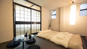 A hotel room with a futon bed, a small round table with a vase and a bottle, a sliding shoji screen door, and a hanging light fixture.