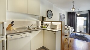 Eine kompakte Küche mit modernen Geräten, ein Essbereich mit weißem Tisch und Stühlen sowie ein Blick ins Freie durch ein großes Fenster.
