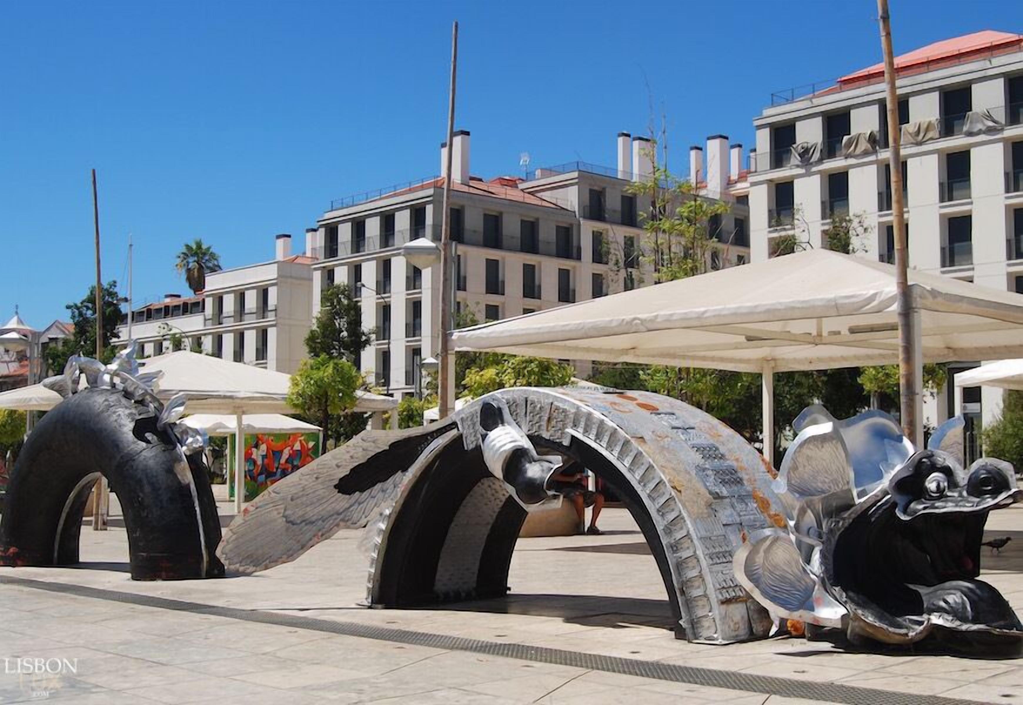 Une sculpture représentant une grande créature mécanique, installée sur une place publique, avec des bâtiments en arrière-plan.