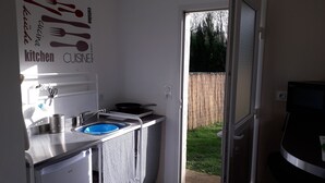 Eine kleine Küche mit Spüle, Herd und Kühlschrank. Eine Tür führt in einen Garten mit Holzzaun.