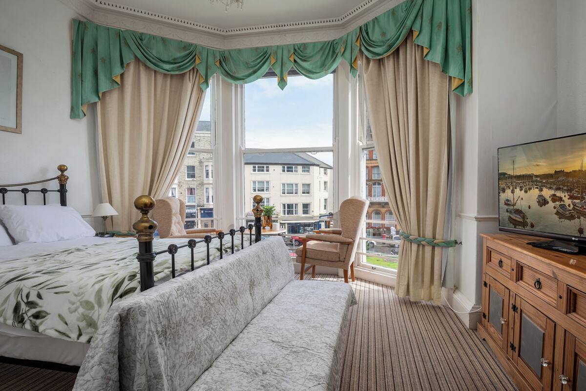 Une chambre d’hôtel équipée d’un lit, d’un canapé, d’une télévision et offrant une vue sur la ville grâce à de grandes fenêtres.