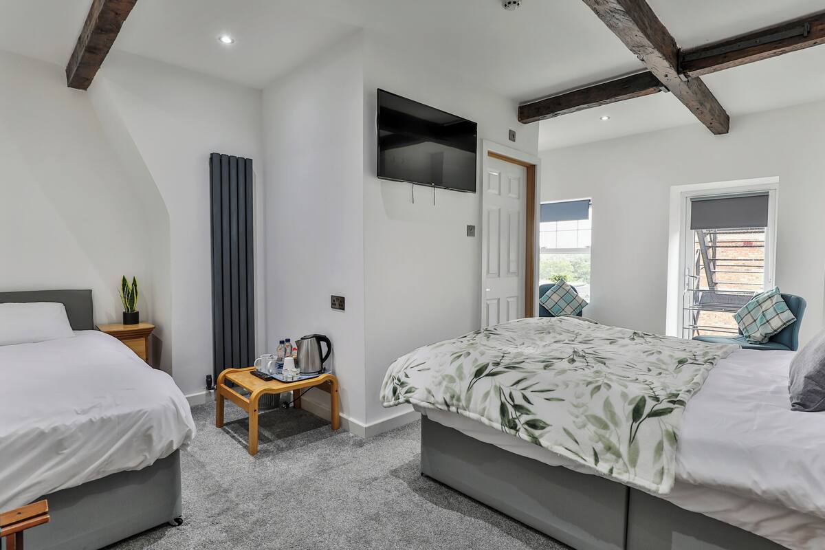 Une chambre avec deux lits, une télévision, un radiateur, une petite table avec une bouilloire et une fenêtre avec des stores.