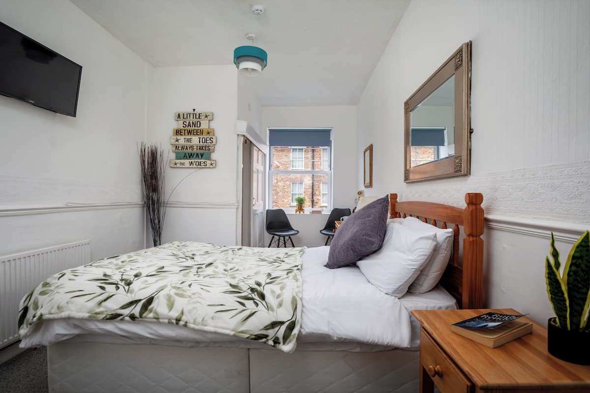 Une chambre à coucher avec un lit, une table de chevet et un miroir. Il y a un panneau sur le mur et une fenêtre donnant sur un immeuble.
