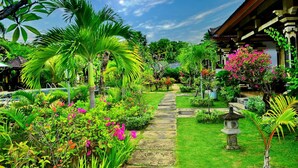 A garden pathway leading to a traditional building with a tiled roof, surrounded by lush greenery and flowering plants.