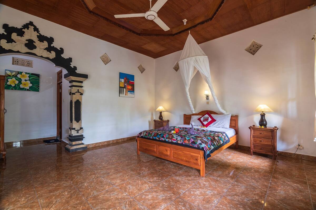 Een slaapkamer met een houten bed, een plafondventilator, een nachtkastje met een lamp en een decoratief wandobject.