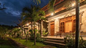 A well-lit traditional building with a stone pathway leading to the entrance, surrounded by lush greenery and palm trees.