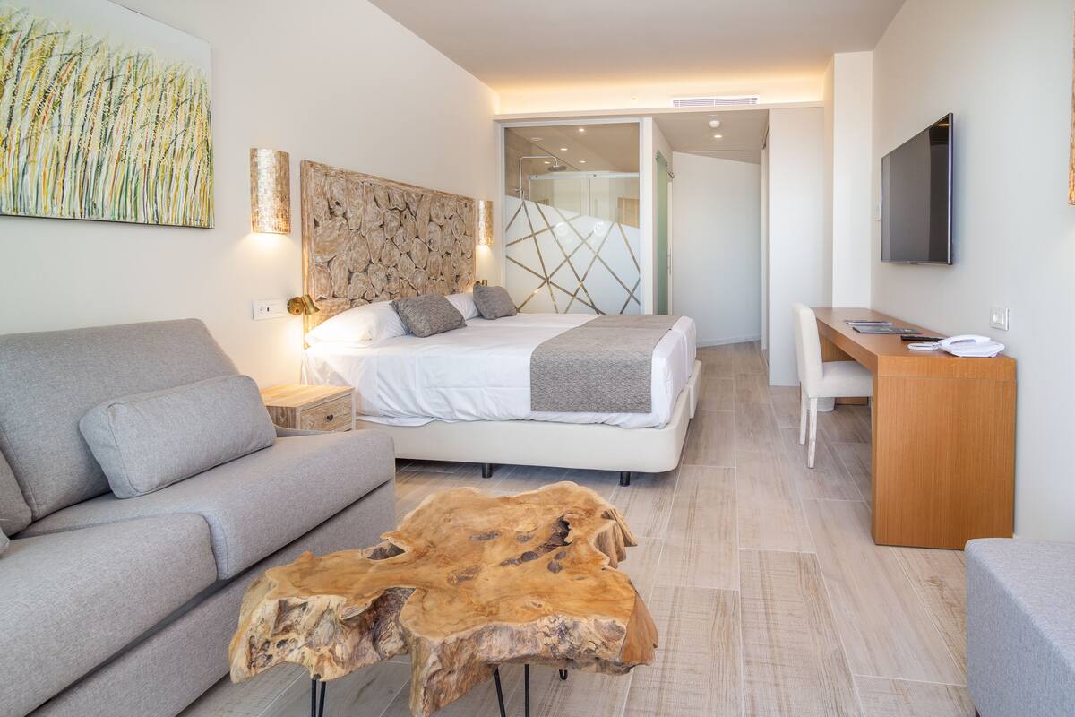 A modern hotel room with a bed, a grey sofa, a wooden coffee table, a desk, and a TV.