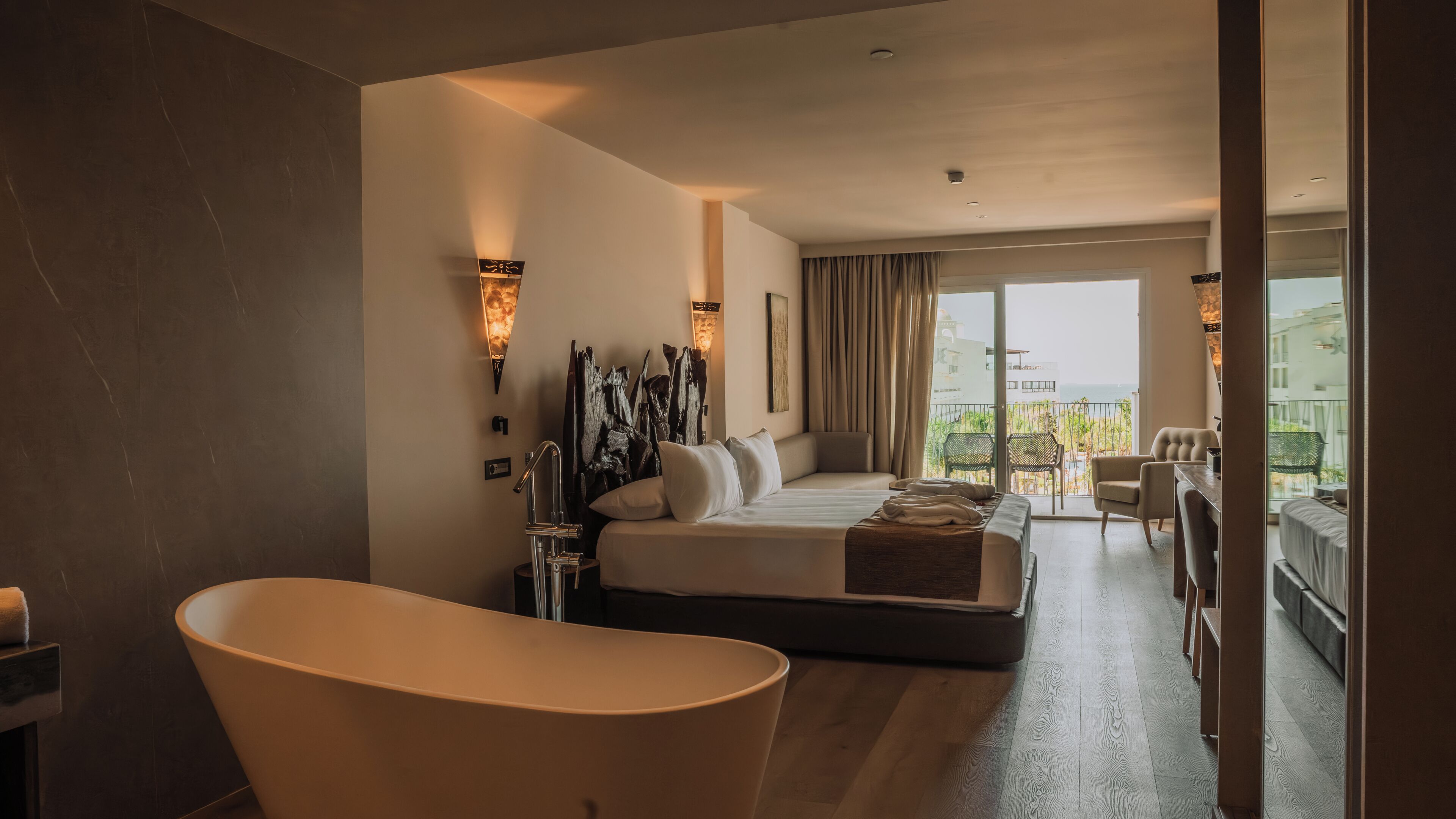 A hotel room with a large bathtub, a bed with a view to the balcony, and a dining area with a view.