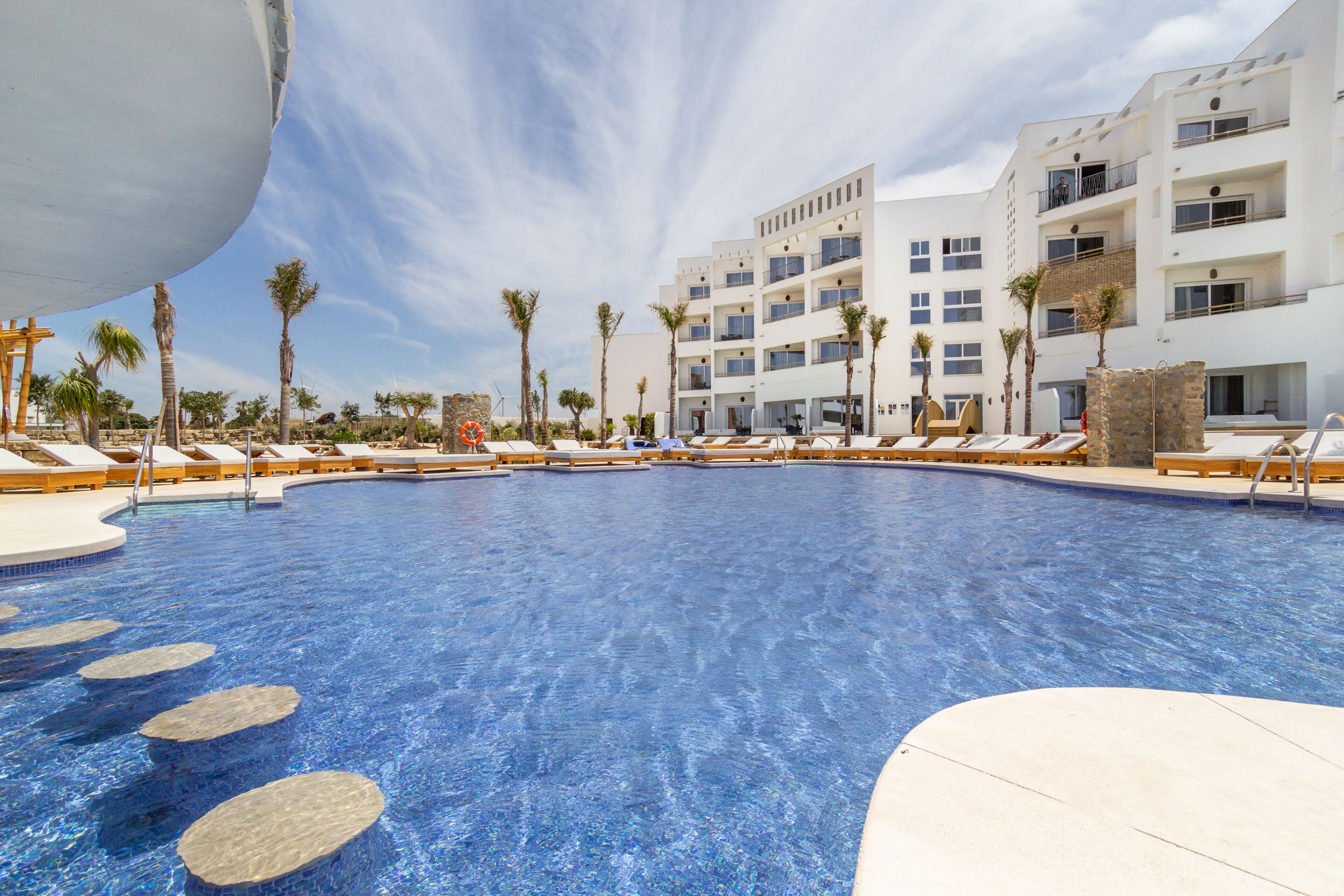 A swimming pool with lounge chairs and palm trees, surrounded by a multi-story building with balconies.