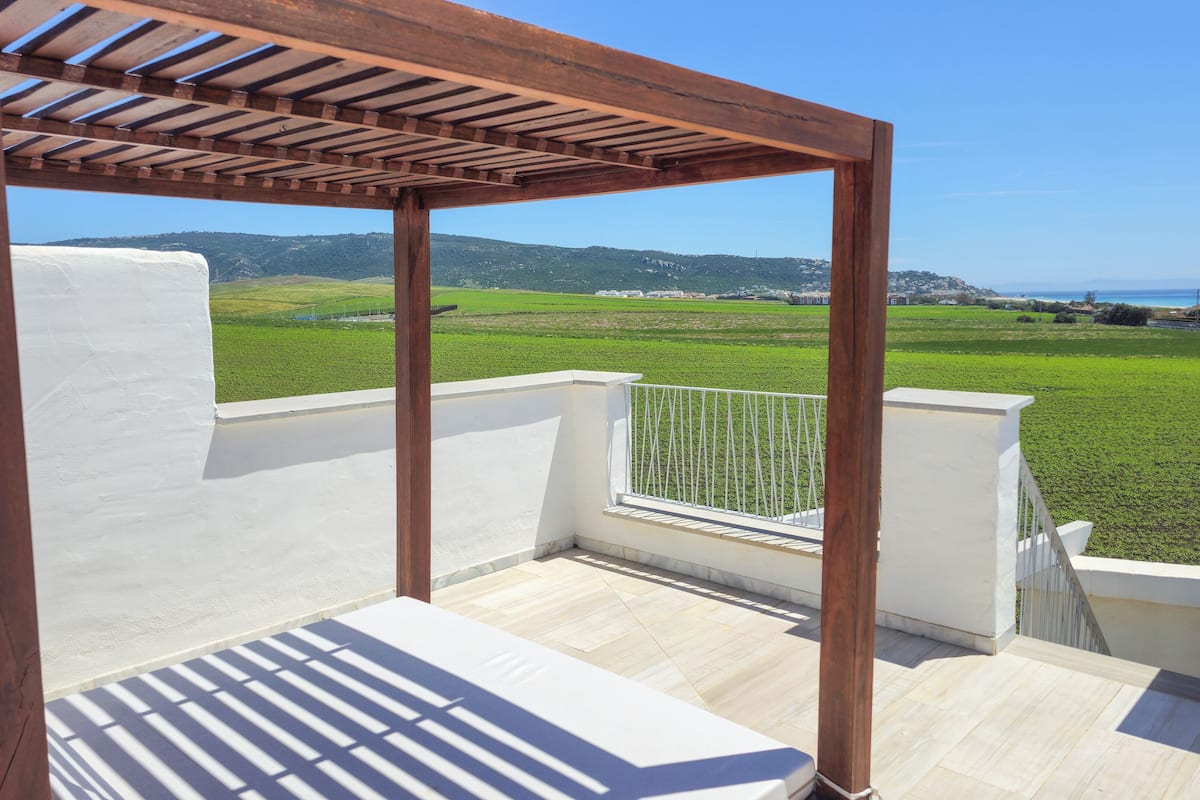 A balcony with a wooden pergola, white walls, and a view of green fields and distant hills.