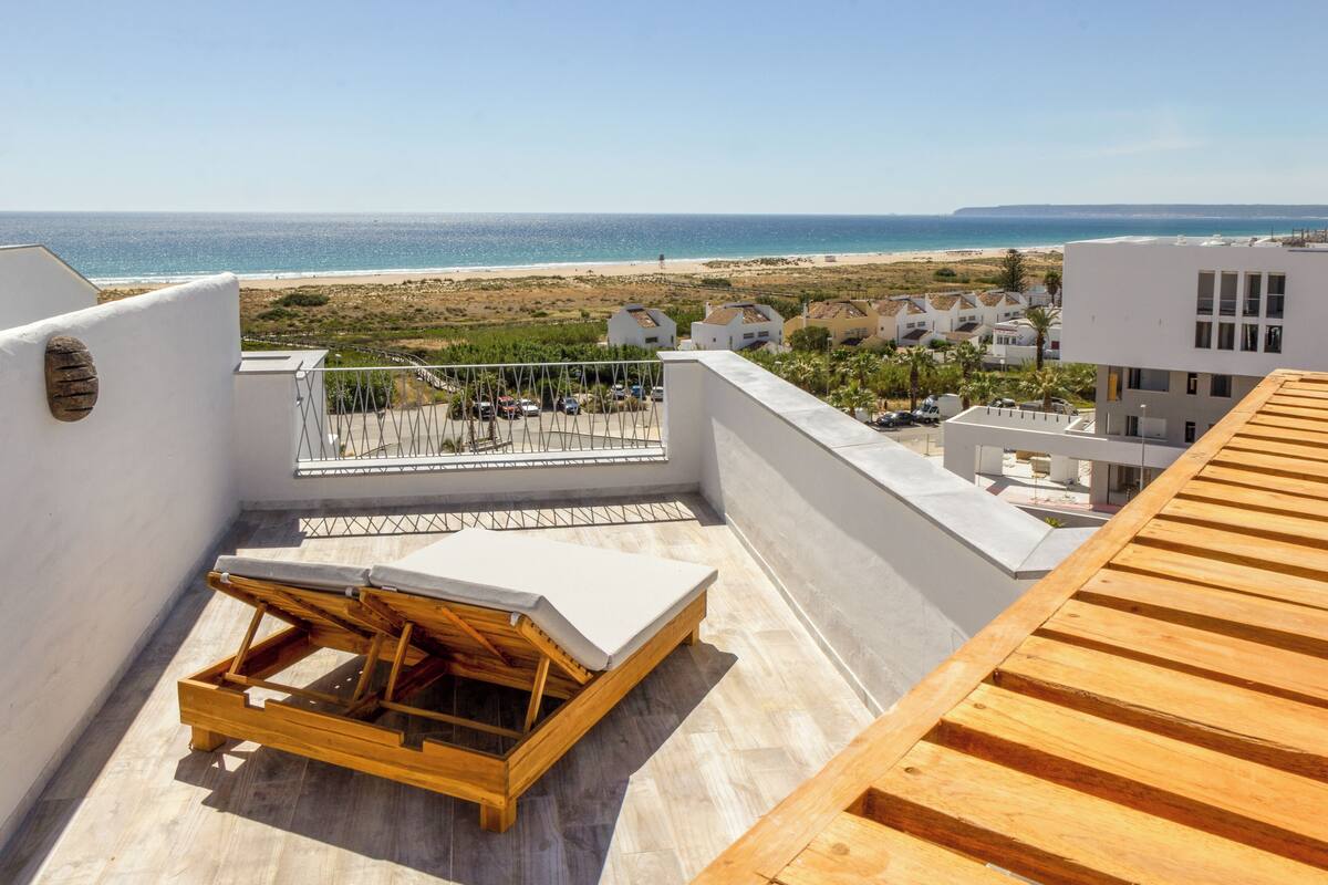 A rooftop terrace with a wooden lounger and a view of the beach and ocean.