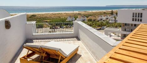 A rooftop terrace with a wooden lounger and a view of the beach and ocean.