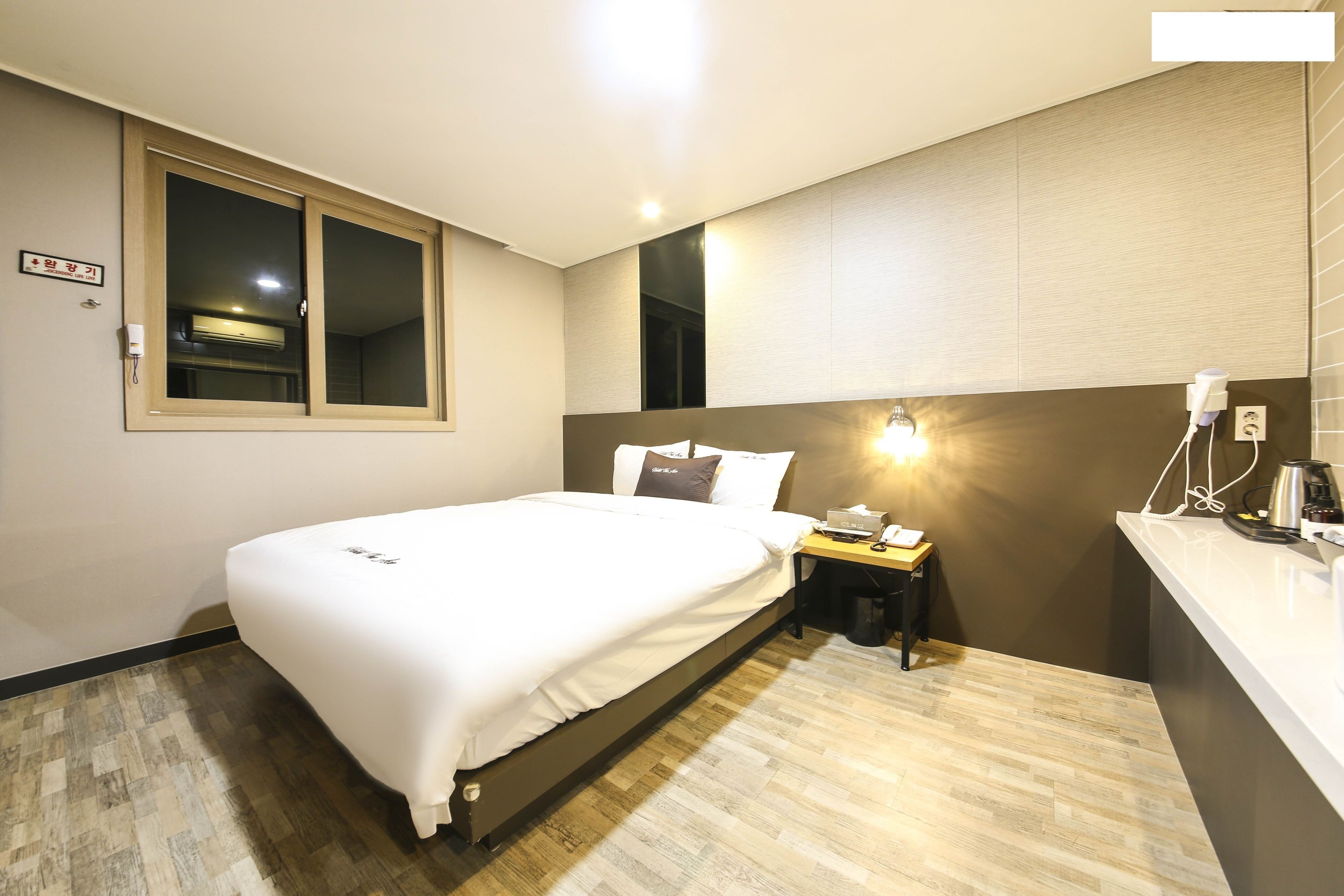 A hotel room with a bed, bedside table, lamp, and a small desk with a chair.