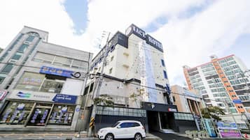 Un hotel con un letrero llamativo, un coche blanco estacionado adelante y varios comercios en la planta baja.