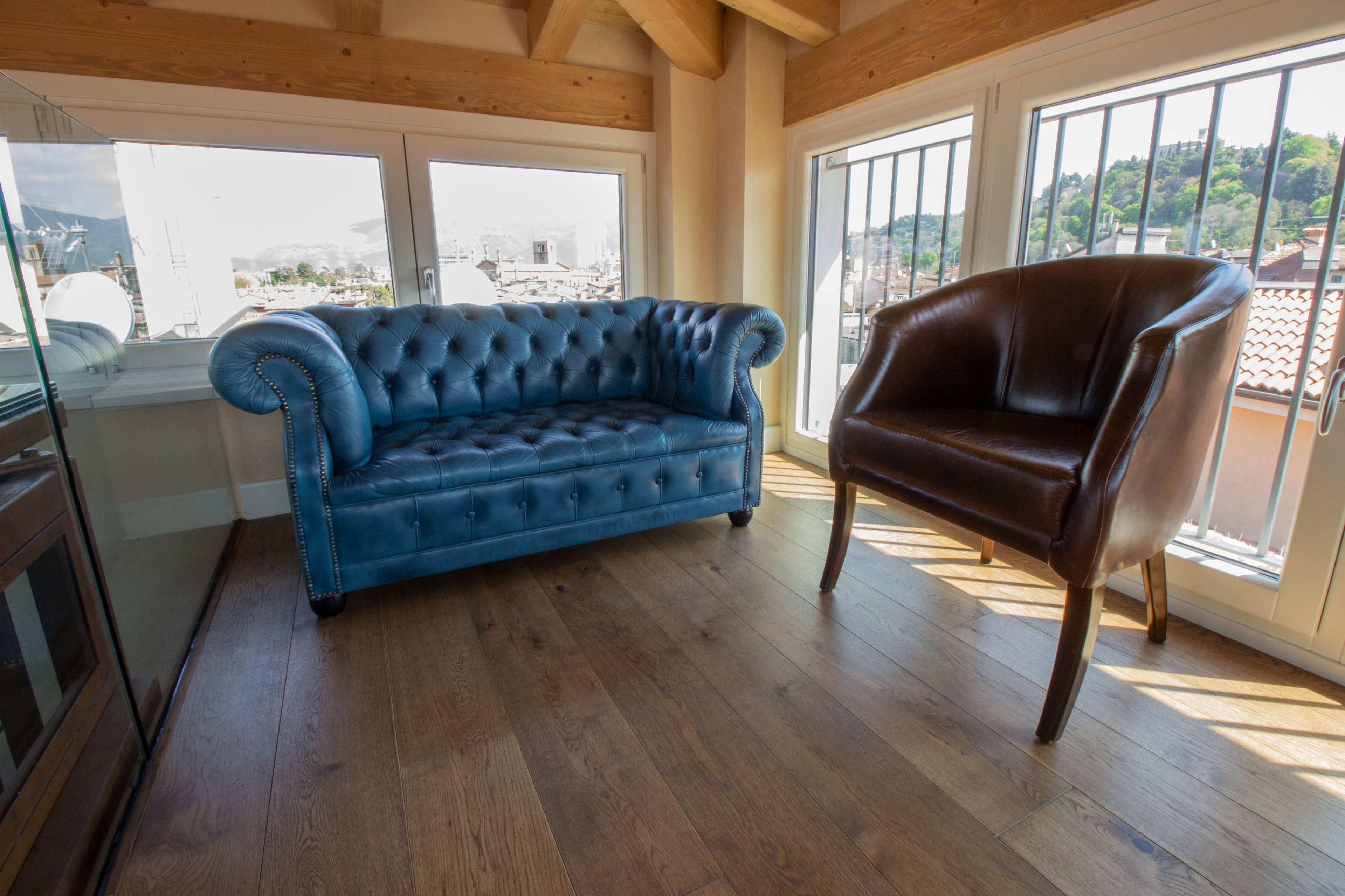 Ein Zimmer mit einem blauen, samtenen Sofa und einem braunen Ledersessel, Holzboden, großen Fenstern mit Stadtblick und einer Holzziel.