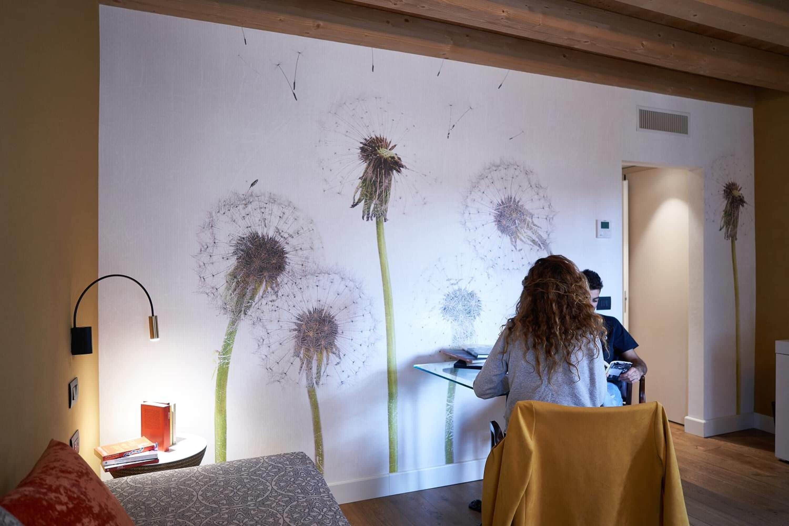 Una habitación con un gran mural en la pared de diente de león, una cama con ropa de cama estampada, un escritorio con una computadora y una lámpara.
