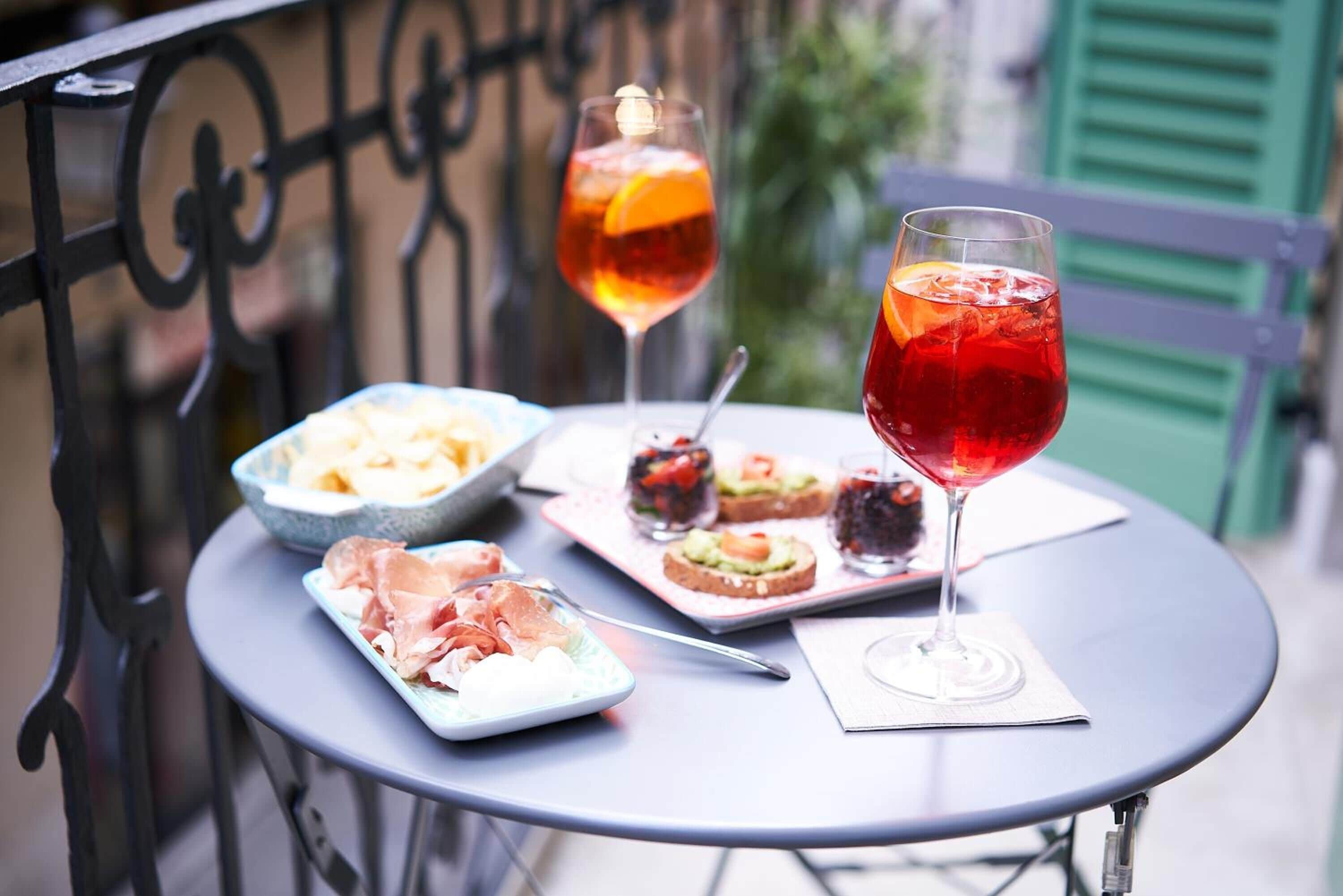 A round table with a white placemat, a glass of red beverage, and a plate of food including prosciutto and bread.