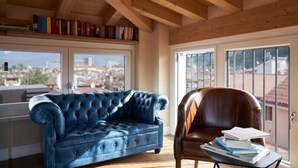 A room with a blue leather sofa, a brown leather armchair, and a table with books. There are large windows with a view of buildings outside. - Locanda delle Mercanzie (Brescia)