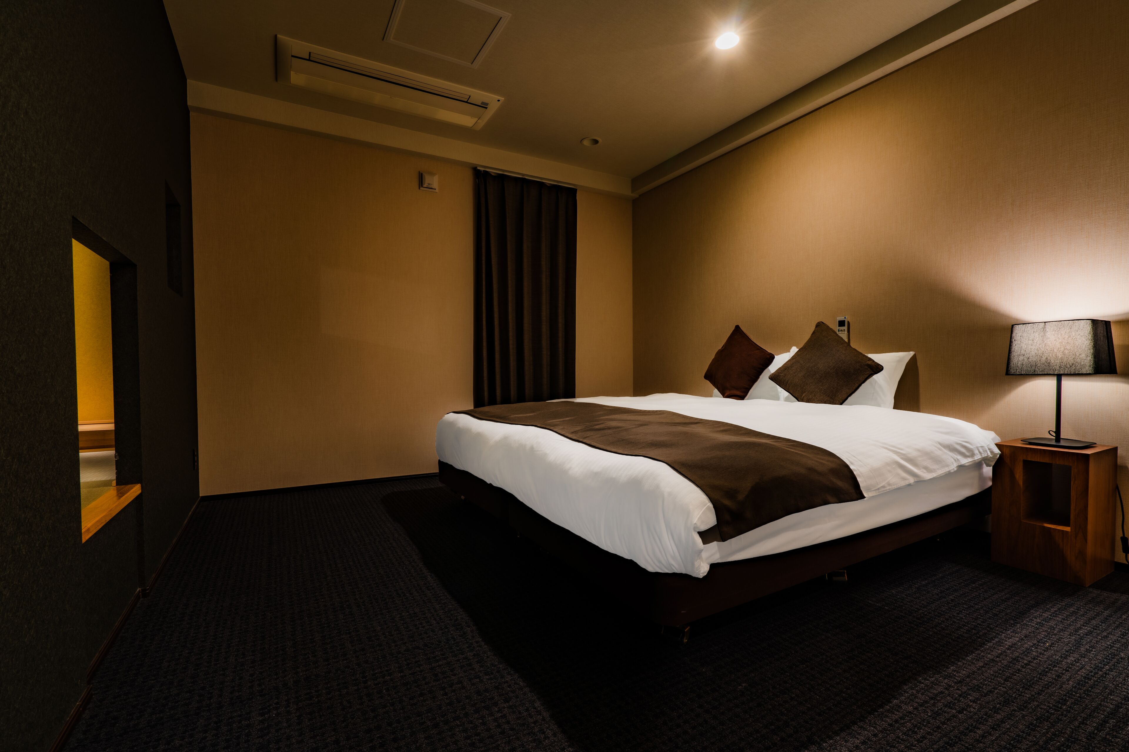 Una habitación de hotel con una cama grande, una mesita de noche con lámpara, una ventana con cortinas y un equipo de aire acondicionado en la pared.