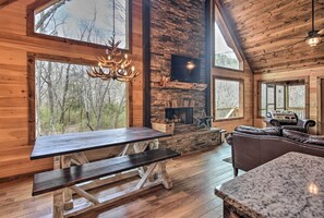 Cozy cabin interior with a high ceiling, stone fireplace, and large windows offering forest views.