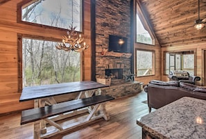 Cozy cabin interior with a high ceiling, stone fireplace, and large windows offering forest views.