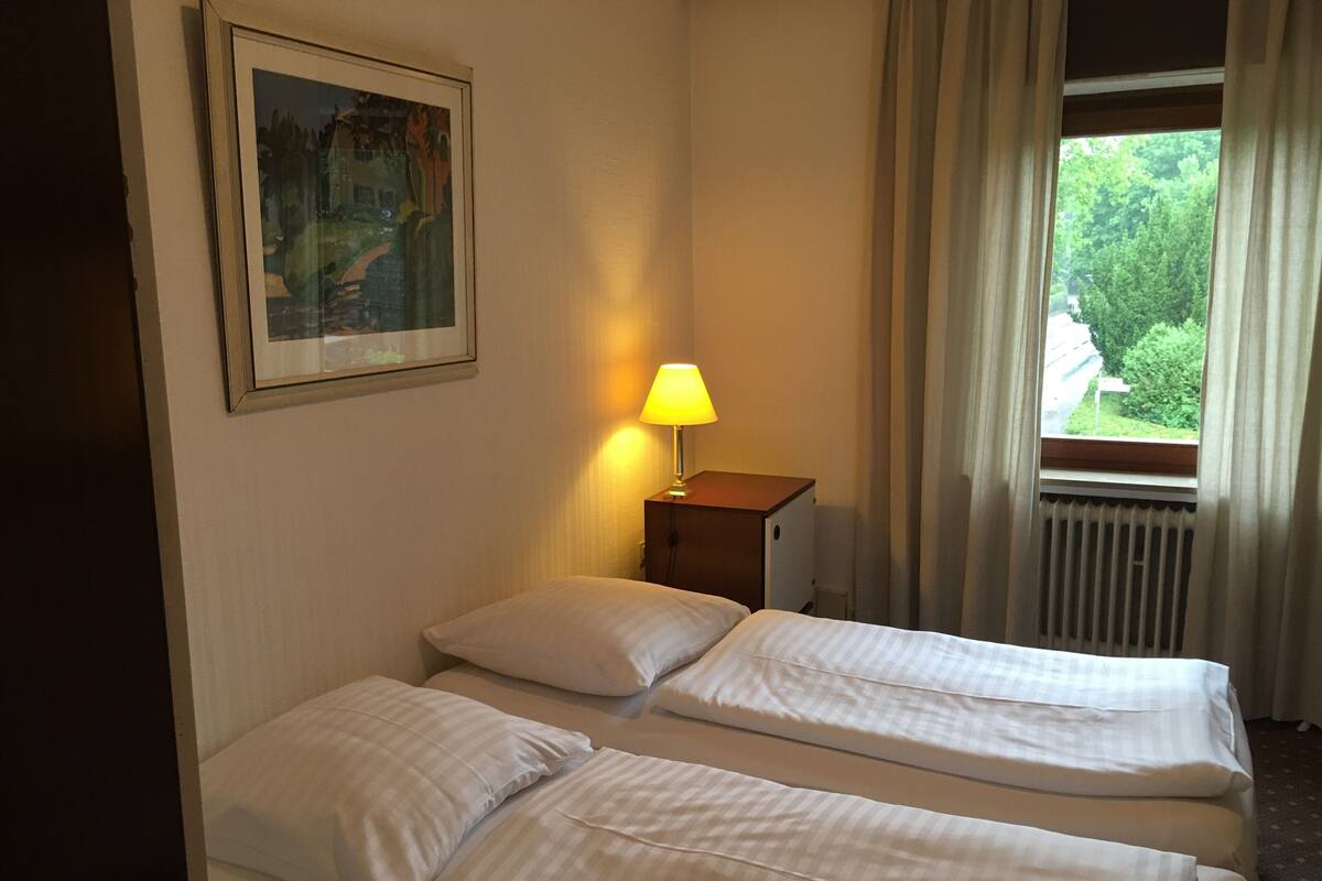 Une chambre d’hôtel avec deux lits, une table de chevet avec une lampe, un tableau au mur et une fenêtre avec des rideaux.
