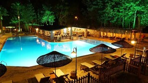 Piscina con sillones de descanso y sombrillas, rodeada de una cerca de madera y árboles.