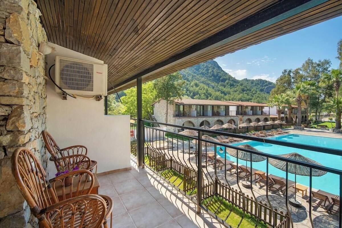 Un balcon avec des fauteuils en osier, une piscine et une vue sur un complexe hôtelier avec ses bâtiments et les montagnes environnantes.