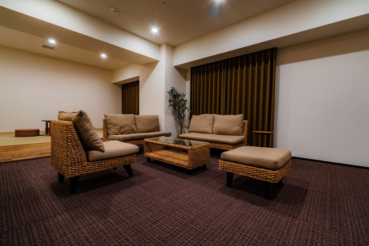 Ein modernes Wohnzimmer mit drei Rattan-Sofas und einem Couchtisch, Holzboden und einem großen Fenster mit Vorhängen.